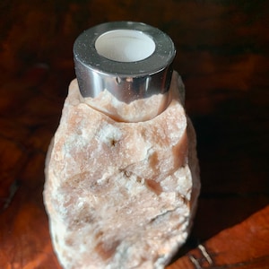 Puede incluir: Un portavelas de metal plateado se asienta sobre una roca natural rosa y rugosa. El portavelas tiene una abertura redonda y lisa para una vela.