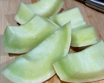 Puede incluir: Seis rebanadas de melón cantalupo verde pálido sobre una tabla de cortar de madera.