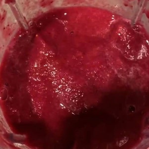 May include: A close-up shot of a blender filled with a vibrant, deep red liquid. The mixture appears thick and smooth, with subtle variations in color and texture, suggesting a freshly made smoothie or juice. The blender's interior is visible.