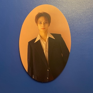 May include: A photo card featuring a man with brown hair wearing a dark blue suit jacket over a white patterned shirt. The background is a light peach color.