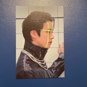 Puede incluir: Una tarjeta de foto con un hombre de pelo negro que lleva una chaqueta de cuero negra con un collar de cadena de plata. El fondo es una pared de azulejos blancos con líneas amarillas.