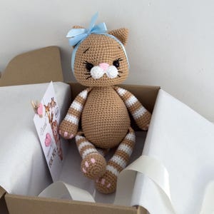 May include: A crocheted tan cat doll with white striped arms and legs, featuring a light blue ribbon bow on its head, black embroidered eyes, and a pink nose. A white card with animal illustrations and the text With sympathy for you is included.