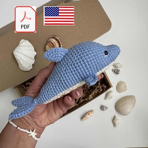 May include: A light blue crocheted dolphin toy with a white belly, fins, and tail. The dolphin is approximately 8 inches long. The image includes seashells, a PDF icon, and an American flag, suggesting a nautical or ocean-themed product.