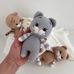 May include: Three handmade crochet cat dolls featuring stitched facial details, including black eyes, pink noses, and white muzzles with whiskers. One doll is grey with striped limbs, while the other two are tan and cream with brown striped accents.