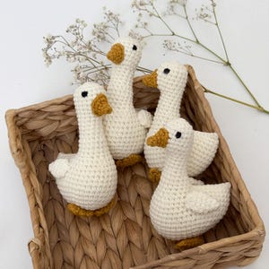 May include: Four handmade crochet geese in a woven basket. The geese are white with yellow beaks and feet, and black eyes. The basket is a light brown color. A sprig of white flowers is in the background.