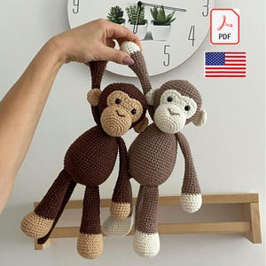 Puede incluir: Dos muñecos de mono de ganchillo, marrón y beige. Uno cuelga del brazo, el otro está de pie. Los muñecos tienen caras detalladas y probablemente sean juguetes u objetos decorativos. La imagen también muestra un reloj.