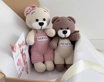 May include: Two handmade crochet teddy bears, one beige and one brown, wearing overalls. The beige bear has a pink bow and pink overalls with the name "MAYA". The brown bear has beige overalls with the name "HENRY". A card with the words "With especially for you" is included.