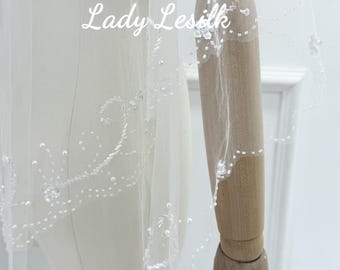 May include: A white bridal veil with intricate beaded detailing along the edge. The veil is draped over a mannequin, highlighting its flowing fabric and delicate design. The text "Lady Lesilk" is visible on the veil.