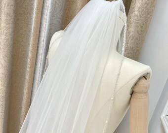 May include: A white bridal veil with a single row of pearl beading along the edge. The veil is draped over a dress form.