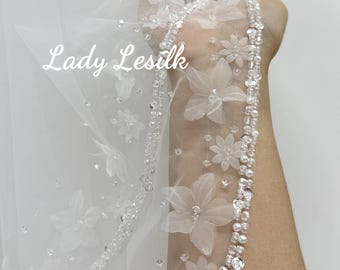 May include: A sheer white bridal veil with delicate floral appliqués and sparkling bead accents. The edge is adorned with a row of clear beads and small, fabric flowers. The text "Lady Lesilk" is visible on the veil.