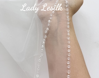 May include: A delicate white bridal veil with a beaded trim and scattered beads. The text "Lady Lesilk" is visible on the veil. The veil is held up against a white background.