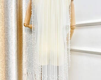May include: A white bridal veil with a single layer of tulle and pearl embellishments. The veil is draped over a mannequin and has a scalloped edge.