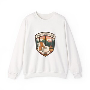 Peut inclure: Sweat-shirt blanc avec un motif graphique d'une scène intérieure confortable. Le motif représente un fauteuil inclinable avec une couverture, une table d'appoint et une vue par la fenêtre sur un paysage de montagne. Le texte "INDOORSY NATIONAL PARK" et "Est. Staying In" sont également inclus.