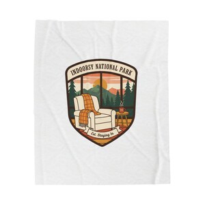 Könnte beinhalten: Weiße Decke mit einer Grafik einer gemütlichen Szene. Die Grafik zeigt einen Blick aus dem Fenster auf eine Berglandschaft, einen Sessel mit einer karierten Decke und eine Kaffeetasse. Der Text lautet "Indoorsy National Park" und "Est. Staying In."