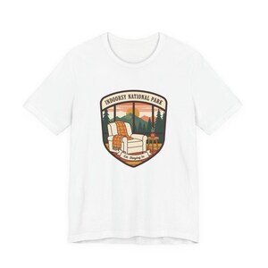 May include: White t-shirt with a graphic depicting an armchair inside a window with a mountain view. The graphic includes the text "Indoorsy National Park" and "Est. Staying In."