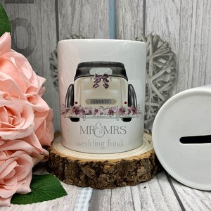 May include: A white ceramic money bank with a wedding car design. The bank features a vintage car illustration with floral accents and the text "MR & MRS wedding fund". The bank is cylindrical and sits on a wooden slice.
