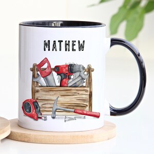 May include: White ceramic mug with a black rim and handle. The mug features a black and white graphic of a toolbox with tools, including a hammer, tape measure, saw, and drill. The name "MATHEW" is printed in black above the toolbox.