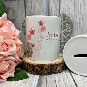 May include: White ceramic money box with a slot on the top. The box is decorated with pink and brown illustrations of a teddy bear, balloons, and blocks that spell out "BABY". The text "Mia Martinez" is written on the box.