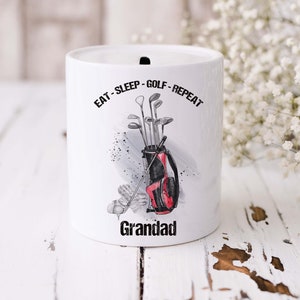 May include: White ceramic money box with a golf bag and clubs design. The text "Eat - Sleep - Golf - Repeat" is printed on the mug. The word "Grandad" is printed at the bottom of the mug.