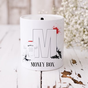 May include: White ceramic money box featuring a black and white football shirt, black and white football boots, and a large black and white letter M. The text "MONEY BOX" is printed on the front of the box.