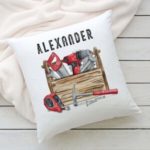 May include: White square cushion featuring a watercolour illustration of a wooden toolbox filled with tools. The toolbox contains a saw, drill, wrench, hammer, tape measure, and nails. The name "ALEXANDER" is printed in black above.