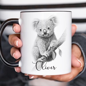 May include: White ceramic mug with a black handle. The mug features a black and white pencil drawing of a koala bear sitting on a branch. The name "Oliver" is written in cursive below the koala.