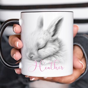 May include: White ceramic mug with a black handle. The mug features a black and white illustration of a sleeping bunny with the name "Heather" written in pink cursive.