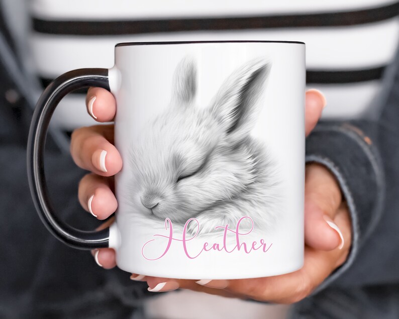May include: White ceramic mug with a black handle. The mug features a black and white illustration of a sleeping bunny with the name "Heather" written in pink cursive.