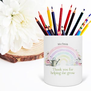 May include: A white pencil holder with a rainbow design, butterflies, and a watering can. The text reads "Thank you for helping me grow" and "Mrs Smith." The holder is filled with various colored pencils.