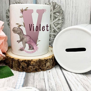 May include: A white ceramic money box with a pink dinosaur and the letter V on the front. The name Violet is written below the letter. The money box is sitting on a wooden log.
