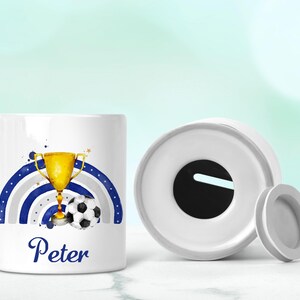 May include: White ceramic money box with a blue and grey rainbow design, a gold trophy, and a football. The name "Peter" is written in blue below the design.
