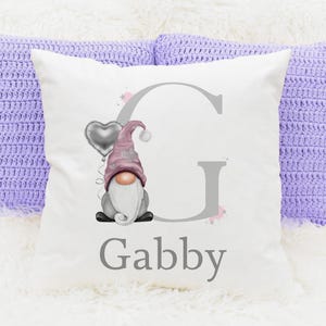 May include: A white cushion featuring a gnome illustration with a pink hat and a silver heart balloon. The letter "G" and the name "Gabby" are printed in grey. Two lilac crochet cushions are in the background.