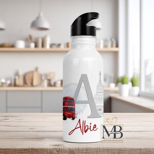 May include: A white water bottle with a black lid featuring a large gray letter A, the name Albie in red script, a red double-decker bus, a gray Ferris wheel, and a gray clock tower illustration. The bottle has a smooth finish and cylindrical shape.