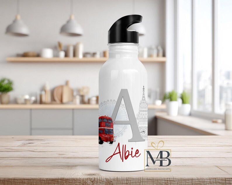May include: A white water bottle with a black lid featuring a large gray letter A, the name Albie in red script, a red double-decker bus, a gray Ferris wheel, and a gray clock tower illustration. The bottle has a smooth finish and cylindrical shape.