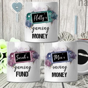 May include: Three white ceramic money boxes with floral designs and a video game controller graphic. The money boxes have the text "Holly's gaming money", "Sarah's gaming fund", and "Mia's saving money".