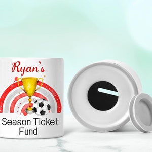 May include: White ceramic money box with a slot on the top. The front of the box has a red and white rainbow design with a gold trophy and a football. The text "Ryan's Season Ticket Fund" is printed on the front of the box.