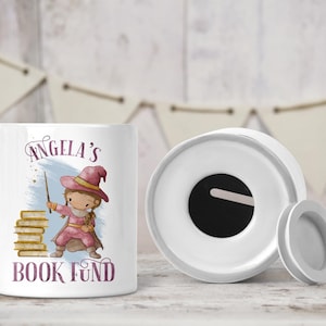 May include: White ceramic money box with a removable lid. The box features a colourful illustration of a girl dressed as a witch with a stack of books and the text "Angela's Book Fund".
