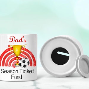 May include: White ceramic money box with a red and white striped design, a gold trophy, and a football. The text "Dad's Season Ticket Fund" is printed on the front.