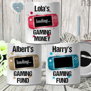 May include: Three white ceramic money boxes with colourful gaming console designs. The money boxes have the names "Lola's", "Albert's", and "Harry's" printed on them. Each money box has the text "loading..." and "GAMING" printed on them. The money box labelled "Lola's" has the text "MONEY" printed on it. The money boxes labelled "Albert's" and "Harry's" have the text "FUND" printed on them.