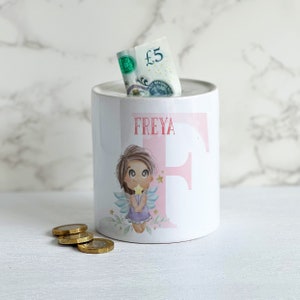 May include: White ceramic money box with a pink and white fairy design. The fairy is holding a star and has wings. The money box has the name "FREYA" written on it.