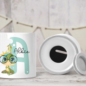 May include: White ceramic money box with a slot on the top. The box features a green cartoon lizard wearing glasses and a crown, sitting next to the letter A. The name "Albie" is written on the box.