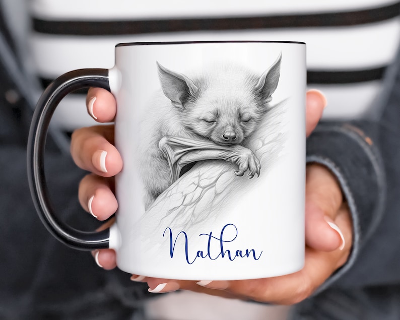 May include: White ceramic mug with a black handle. The mug features a black and white pencil drawing of a bat sleeping on a branch. The name "Nathan" is written in blue cursive below the image.