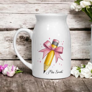 May include: White ceramic mug featuring a yellow pencil design with a pink bow and the text "Miss Smith". The mug has a handle and is suitable for hot or cold beverages.