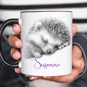 May include: White ceramic mug with a black handle. The mug features a black and white illustration of a sleeping hedgehog. The text "Susanne" is printed in purple below the hedgehog.