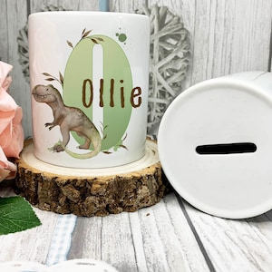 May include: White ceramic money box featuring a dinosaur illustration and the name "Ollie". The money box has a slot for coins and is displayed on a wooden slice. The design includes green and brown accents.