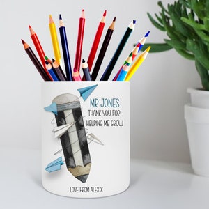 May include: White ceramic pencil holder with a black and white pencil illustration and blue paper airplanes. The text reads "Mr. Jones Thank you for helping me grow Love from Alex X".