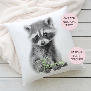 May include: White pillow with a black and white illustration of a raccoon. The raccoon is sitting up and looking at the camera. The text "Thomas" is written in green cursive below the raccoon. The text "Can add your own text" is in a pink circle in the top right corner of the pillow. The text "Various font colours" is in a pink circle in the bottom right corner of the pillow.