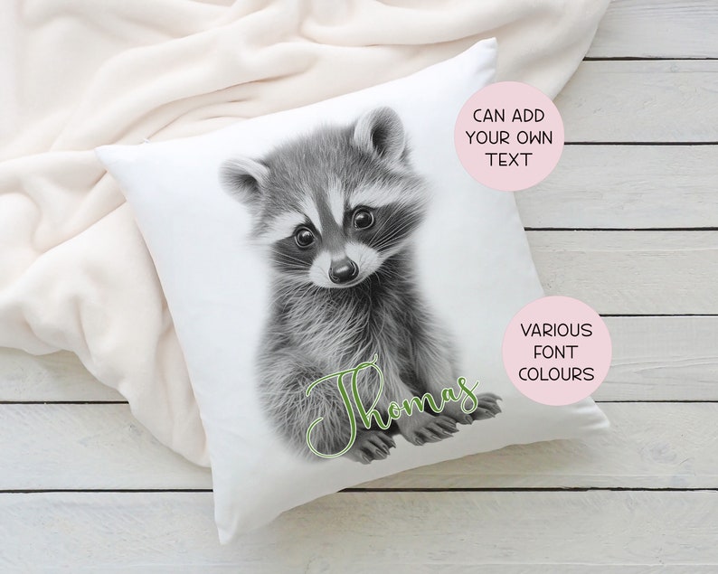 May include: White pillow with a black and white illustration of a raccoon. The raccoon is sitting up and looking at the camera. The text "Thomas" is written in green cursive below the raccoon. The text "Can add your own text" is in a pink circle in the top right corner of the pillow. The text "Various font colours" is in a pink circle in the bottom right corner of the pillow.