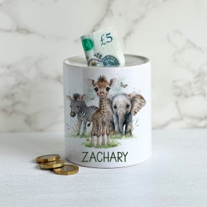 May include: White ceramic money box featuring a giraffe, zebra, and elephant illustration. The money box has the name "ZACHARY" printed on the front.