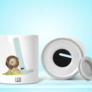 May include: White ceramic piggy bank with a lion illustration and the letter L. The lid is off and the bank has a slot for coins. The name Leo is printed on the front of the bank.